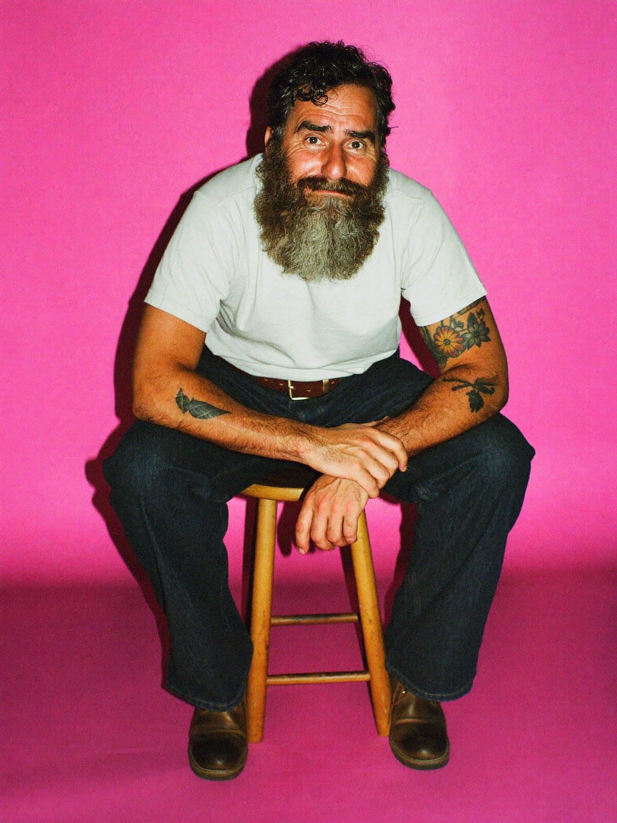 A bearded man seated on a stool against a pink backdrop
