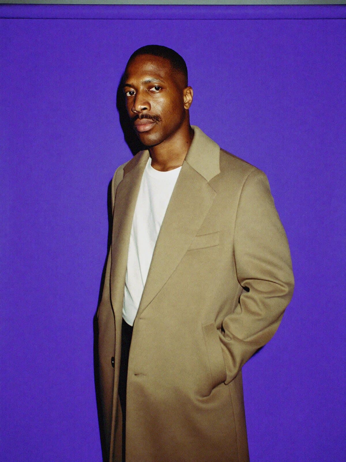 A man in a camel overcoat against a violet backdrop